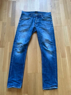 Blå raka jeans från Tiger of Sweden - Säljer ett par blå jeans från Tiger of Sweden med raka ben och klassisk femficksmodell. Jeansen har slitningar och tvättade detaljer för en cool, använd look. Tillverkade i denim med kontrastsömmar och knappgylf. Litet hål vid skrevet.