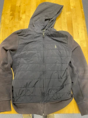 Polo Ralph Lauren cardigan - Snygg svart pufferjacka från Polo Ralph Lauren med broderad logga på bröstet. Jackan har huva, dragkedja framtill och quiltad framsida i glansigt material medan ärmarna är i mjuk fleece. 