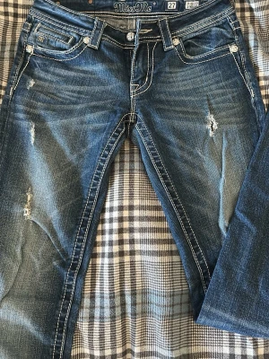 Blå bootcut jeans från Miss Me - Säljer ett par blå bootcut jeans från Miss Me med slitningar och kontrastsömmar. Jeansen har fem fickor, dekorativa nitar och en klassisk låg midja. Materialet är denim i bomull med lite stretch för skön passform.