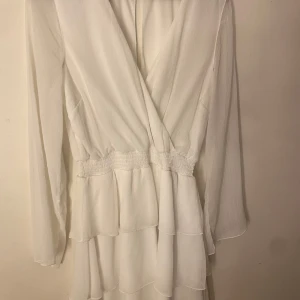 Vit omlottklänning från Bubbleroom - Elegant vit omlottklänning från Bubbleroom med långa ärmar och smockdetaljer i midjan. Klänningen har en v-ringad design och ett flödande tyg som ger en luftig känsla. Endast använd 1 gång. 