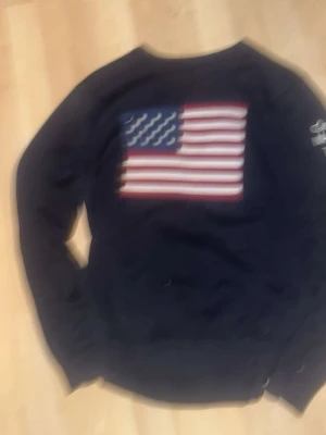Svart sweatshirt med USA-flagga - Cool svart sweatshirt med stor amerikansk flagga tryckt på ryggen. Tröjan har rund hals och långa ärmar, perfekt för en avslappnad streetstil. Passar dig som gillar klassiska och sportiga plagg med en touch av USA.