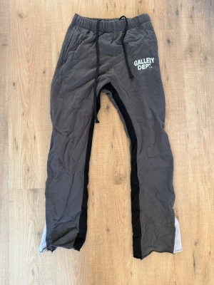 Grå mjukisbyxor från Gallery Dept. - Snygga grå mjukisbyxor från Gallery Dept. med svart och vit panel längs benen. Byxorna har resår i midjan med dragsko och tryckt logga på vänster lår. Perfekta för en chill och avslappnad stil. Pris kan diskuteras vid snabb affär.