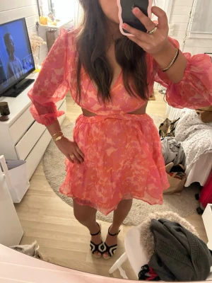 Rosa blommig kortklänning med volang - Superfin kort klänning i rosa och orange toner med blommigt mönster och volangkant. Djup v-ringning, markerad midja och långa puffärmar i transparent material. Perfekt för sommarens alla tillfällen! Har aldrig använt den själv.