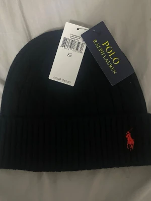 Svart mössa från Polo Ralph Lauren - Svart ribbstickad mössa från Polo Ralph Lauren med klassisk röd broderad logga framtill. Mössan har uppvikt kant och är tillverkad i mjukt material som håller dig varm. Perfekt accessoar för kyliga dagar.