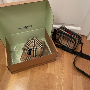 Burberry Keps & Väska (Combo) - Säljer en snygg rutig keps från Burberry i klassiskt beige, svart och rött mönster. Kepsen har en justerbar rem baktill och ett broderat monogram på framsidan. Perfekt för att ge din outfit en lyxig touch. Keps skick 8/10 går att tvätta, väska skick 8/10