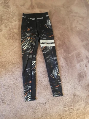 Mönstrade leggings från Stronger - Snygga svarta leggings från Stronger med färgglatt abstrakt mönster i orange, vitt och grått. Hög midja med logotyp i midjebandet och två vita ränder på ena benet. Perfekta för träning eller chill. Stretchigt material för skön passform.
