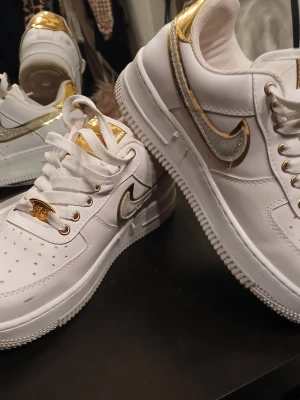 Nike vita sneakers med guld och silver - Snygga vita Nike sneakers med metallic-detaljer i guld och silver. Skorna har klassisk låg modell, vit sula och perforerad tå. Guldiga detaljer på häl, snörning och logga ger en riktigt fräsch och trendig look. Perfekta för dig som vill sticka ut lite extra.