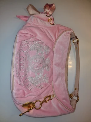 Juicy Couture rosa velour handväska - Söt rosa handväska från Juicy Couture i mjuk velour med broderad logga framtill. Väskan har guldfärgade metalldetaljer, beige axelrem och en rosett upptill. Insidan är fodrad i guld och rosa med tryck och har flera fack, bland annat med texten 'Hello?' och 'SHOP!'.