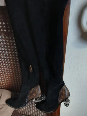 Svarta overknee-stövlar Louis Vuitton - Snygga svarta overknee-stövlar från Louis Vuitton med klassiskt monogrammönster i brunt på hälen och bakre delen. Stövlarna har spetsig tå, dragkedja med detalj och unika klackar med kedjedetaljer. Materialet är mocka och läder.