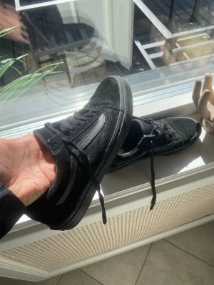 Svarta Vans Old Skool sneakers - Klassiska svarta Vans Old Skool sneakers med lågt skaft. Skorna har ovandel i mocka och canvas, ikonisk sidosnöre i svart och svarta skosnören. Perfekta för en streetwear-look och passar till det mesta i garderoben.