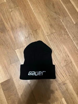 Svart Bauer mössa med brodyr - Säljer en svart mössa från Bauer med vit broderad logga framtill. Klassisk ribbstickad design med uppvikt kant. Perfekt för dig som gillar sportig stil och vill hålla dig varm under kalla dagar.