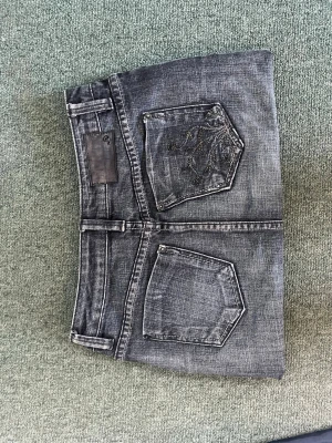Grå jeanskjol med detaljer - Snygg grå/blå jeanskjol med slitna detaljer och dekorativa sömmar. Kjolen har två bakfickor, varav en är dekorerad med svarta paljetter. Klassisk femficksmodell och kort passform, perfekt till vardagslooken.
