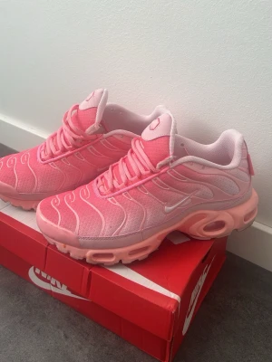 Nike Air Max Plus rosa sneakers - Nike Air Max Plus sneakers i en snygg rosa färg med vita detaljer. Ovandelen är i mesh och syntetmaterial med vågiga linjer och synlig Air Max-dämpning i sulan. Skorna har en sportig vibe och är perfekta för dig som gillar färg och streetstyle. Och den är helt Ny.