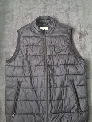 Svart dunväst från Jack & Jones - Stilren svart dunväst från Jack & Jones med quiltad design och hög krage. Västen har två dragkedjeförsedda fickor och en diskret logotyp på bröstet. Perfekt för lager-på-lager och enkel att matcha med olika outfits.