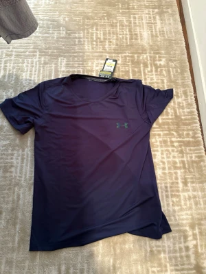Mörkblå tränings-tshirt Under Armour - Snygg mörkblå tränings-tshirt från Under Armour med klassisk logga på bröstet och text längs ryggen. Tillverkad i ett lätt och snabbtorkande funktionsmaterial som passar perfekt till gymmet eller löprundan. Rund halsringning och korta ärmar.