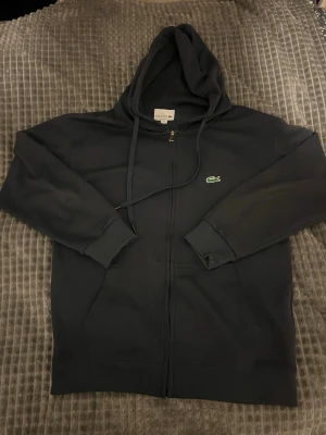 Lacoste Mörkblå ziptröja  - Säljer en mörkblå oanvänt zip hoodie från Lacoste med dragkedja och klassisk krokodillogo på bröstet som jag säljer pga att den blev för liten. Den är i storleken M men passar även i storleken S. Tröjan har huva med snören, ribbade muddar och två fickor framtill. Perfekt för en avslappnad och cool look.