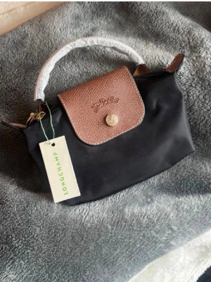 Svart handväska från Longchamp - Snygg svart handväska från Longchamp med brunt lock i präglat läder och guldfärgade detaljer. Väskan har ett kort handtag och dragkedja upptill. Axelrem tillkommer❤️ Helt ny! 