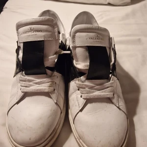Valentino Garavani vita sneakers - Säljer ett par vita Valentino Garavani sneakers med bred svart läderdetalj över plösen. Klassisk rund tå, snörning och robust sula med Valentino-logga undertill. Snygg kontrast mellan vitt och svart, perfekt för dig som gillar exklusiva streetwear-vibbar. Strl är för liten för mig så jag säljer dem vidare . är sköna och ser bra ut