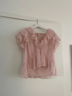 Rosa volangblus med kort ärm - Supersöt ljusrosa blus med volanger längs ärmar och krage. Blusen är tillverkad i ett tunt, transparent tyg som ger en luftig känsla. Knappar framtill och lös passform gör den extra bekväm och trendig. Perfekt för dig som gillar romantisk och feminin stil.