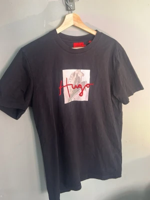 Svart Hugo Boss t-shirt med tryck - Snygg svart t-shirt från Hugo Boss med ett unikt tryck på bröstet och röd broderad 'Hugo'-logga. Klassisk rund hals. Tröjan är S men kan gott och väl passa i lite mindre M. Inga stora defekter men lite sliten är den. Hör av er vid fler frågor!