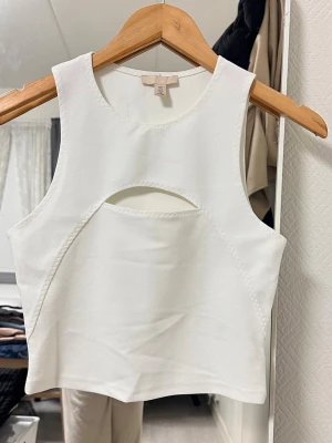 Vit croppad linnetopp med cut out - Trendig vit croppad linnetopp från H&M med rund halsringning och snygg cut out-detalj framtill. Toppen är ärmlös och har en tight passform som framhäver siluetten. Perfekt att styla med jeans eller kjol för en cool look.