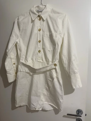 Vit skjortklänning med knappar - Stilren vit skjortklänning med långa ärmar, krage och bröstficka. Klänningen har snygga knappar framtill och en omlott-detalj i kjolen som knäpps i sidan. Perfekt för dig som gillar en clean och trendig look.