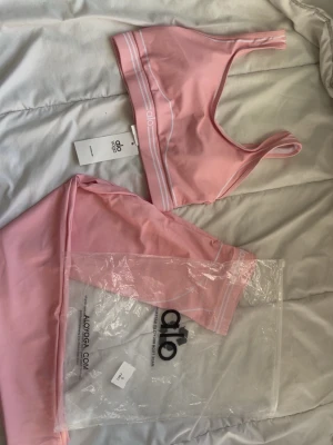 Rosa set från Alo Yoga - Säljer ett snyggt rosa underklädesset från Alo Yoga. Setet består av en sportig topp med smala axelband och matchande trosor, båda med vita detaljer. Materialet är mjukt och stretchigt, perfekt för en bekväm känsla hela dagen.