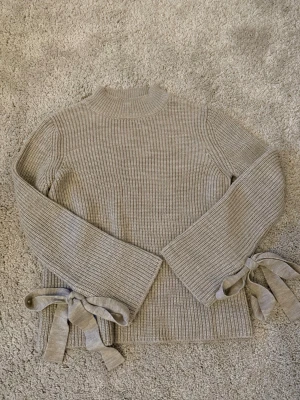 Beige stickad tröja med knytband - Säljer en superfin beige stickad tröja från Gina Tricot, ribbad och med högre hals. Tröjan har långa ärmar med breda muddar och snygga knytband vid ärmsluten.