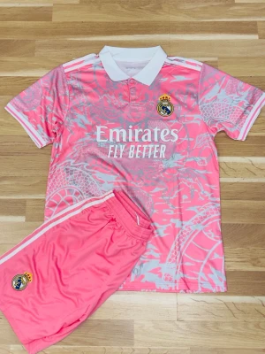 Real Madrid rosa matchställ - Säljer ett rosa matchställ från Real Madrid med vit krage och detaljer. Tröjan har klubbens logga och sponsortryck på bröstet, samt ett unikt mönster i ljusrosa och vitt. Shorts i samma färg med logga och vita ränder på sidorna. Perfekt för fotboll eller träning.