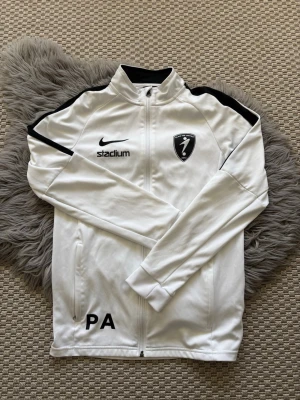 Vit Nike Dri-FIT träningsjacka - Snygg vit träningsjacka från Nike med svarta detaljer och klubbmärke på bröstet. Jackan har hel dragkedja, två fickor och Dri-FIT-material som andas. Märkt med 'PA' och Stadium-logga. Perfekt för träning eller uppvärmning.