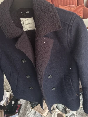 Mörkblå skepparkavaj från Zara - Snygg mörkblå skepparkavaj från Zara med dubbelknäppning och två framfickor. Kragen är fodrad med mjukt teddyfoder för extra värme och stil. Klassisk rak passform och grov ullblandning som ger en tidlös look.