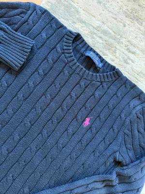 Mörkblå kabelstickad tröja Polo Ralph Lauren - Mörkblå kabelstickad tröja från Polo Ralph Lauren med rund halsringning och ribbade muddar. Tröjan har en rosa broderad logga på bröstet och är tillverkad i ett mjukt stickat material. Perfekt för en stilren och klassisk look.