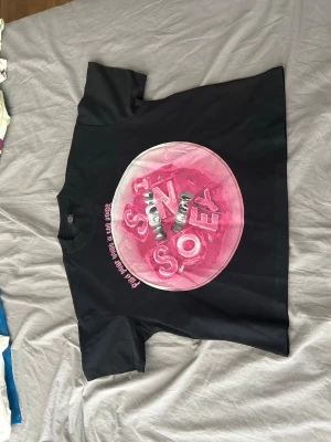 Svart t-shirt med rosa tryck  - Cool svart t-shirt med stort rosa tryck på framsidan som har en rund form och text i metallicstil. T-shirten har klassisk passform och korta ärmar, perfekt för att ge din outfit en färgklick.
