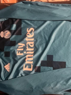 Real Madrid bortatröja Ronaldo Adidas blå - Snygg blå bortatröja från Real Madrid med Fly Emirates-tryck på bröstet och klubbmärke. Tillverkad i lätt och ventilerande material, perfekt för fotboll eller träning. Stora vita siffran 7 på ryggen med namnet Ronaldo och Adidas-logga på framsidan.