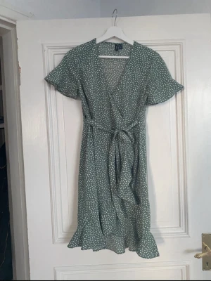 Grön prickig omlottklänning Vero Moda - Säljer en grön omlottklänning från Vero Moda med vita prickar, knytband i midjan och volangkant nertill. Klänningen har korta ärmar och v-ringad hals. Perfekt för dig som gillar en feminin och avslappnad stil.