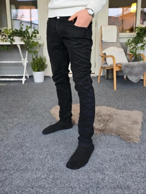 Svarta slim fit jeans herr - Svarta slim fit jeans med klassisk femficksdesign och normal midja. Jeansen har smal passform hela vägen ner och är tillverkade i ett stretchigt material för extra komfort. Perfekta för en stilren och modern look.
