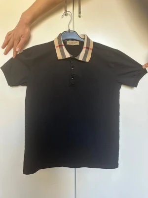 Svart pikétröja från Burberry - Svart pikétröja från Burberry med klassisk krage i beige med röd och svart rutig detalj. Tröjan har korta ärmar och knäppning framtill med flera knappar. Perfekt för en stilren och trendig look. OBS! Storlek M men passar som S.