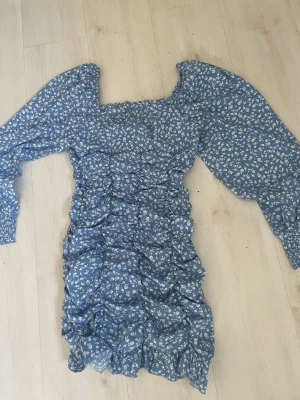 Blå blommig kortklänning med puffärm - Supersöt blå kortklänning med små vita blommor och puffiga långärmar. Klänningen har smockad överdel och rynkade detaljer längs hela plagget för en trendig look. Perfekt för sommarens alla tillfällen!