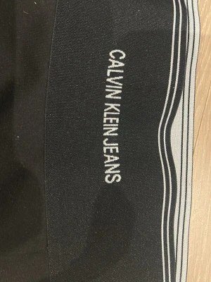Svart kortkjol från Calvin Klein Jeans - Snygg svart kortkjol från Calvin Klein Jeans med bred midjeresår och vit logga framtill. Kjolen har en enkel, tight passform och diskreta vita ränder upptill. Perfekt för en trendig och sportig look.