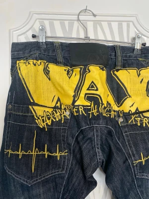 Wax Actionwear baggy jeans med tryck - Säljer ett par mörkblå baggy jeans från Wax Actionwear med stora gula tryck och broderier på baksidan. Jeansen har slitna detaljer, vida ben och coola sömmar. Materialet är klassisk denim och de har en streetwear vibe som sticker ut. Skulle säga att de passar som S-M