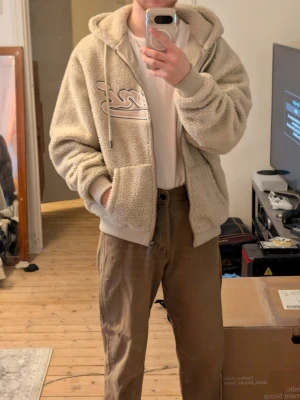 Karl Landy Beige teddy hoodie/jacka med tryck - Säljer en beige teddy hoodie med dragkedja och stor huva. Hoodien har ett broderat tryck på bröstet och breda muddar vid ärmar och nederkant. Materialet är fluffigt och mjukt, perfekt för dig som gillar en avslappnad och cozy stil. Som ny.