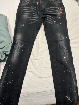 Svarta Dsquared2 jeans  - Säljer ett par svarta jeans från Dsquared2 med coola färgstänk i vitt och orange samt slitna detaljer. Jeansen har raka ben, dragkedja fram och en orange siffra 2 på fickan. Bakfickor med Dsquared2-lapp. Perfekta för en edgy streetstil.