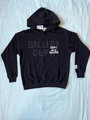 Svart hoodie från Gallery Dept. - Svart hoodie från Gallery Dept. med trycket 'GALLERY DEPT.' i svart och vitt på bröstet. Klassisk känguruficka framtill, justerbar huva med snören och ribbade muddar. Perfekt för en avslappnad och trendig streetwear-look.