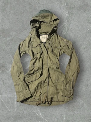 Olivgrön parkas från Abercrombie & Fitch | XS/34 - Stilren olivgrön parkas från Abercrombie & Fitch med flera praktiska fickor, dragsko i midjan och huva. Jackan har en klassisk militärinspirerad look och är tillverkad i slitstark bomull. Perfekt för dig som gillar en avslappnad men trendig stil.