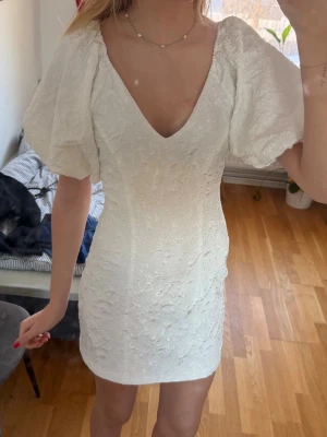 Vit kort klänning med puffärmar - Supersöt vit kort klänning med puffiga kortärmar och v-ringning. Klänningen har ett diskret blommigt mönster i tyget och figurnära passform. Perfekt för sommarens alla tillfällen. Fungerar utmärkt som studentklänning. Endast använd 1 gång.