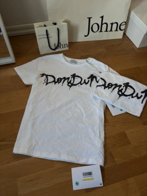 Dondup tshirt med allt og vit - Säljer en helt ny vit t-shirt från Dondup med allt og och ett stort svart graffiti-inspirerat tryck över bröstet. Klassisk rund hals och korta ärmar. Tillverkad i mjuk bomull som känns skön mot huden. Armhåla till armhåla 49cm. Axeln ner 67cm.