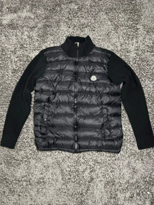 Svart moncler cardigan  - Snygg svart dunväst från Moncler med stickade svarta ärmar och hög krage. Västen har två fickor med knapp framtill och ett klassiskt Moncler-märke på bröstet. Materialet är en kombination av dun och stickad textil, perfekt för lager-på-lager-stil.
