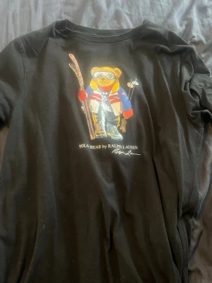Svart Polo Bear t-shirt från Ralph Lauren - Svart t-shirt från Ralph Lauren med ikoniskt Polo Bear-tryck framtill där björnen har skidutrustning och färgglada kläder. Klassisk rund hals och korta ärmar. Det är barn storlek XL men den passar dig som har Xs och kanske S. Köpt ifrån kidsbrandstore 2021