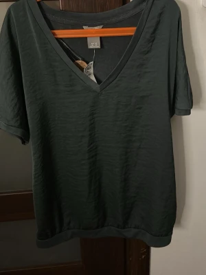 Mörkgrön v-ringad topp från Lindex  - En enkel och stilren mörkgrön topp från Lindex med v-ringning och korta ärmar. Toppen har en loose passform och är tillverkad i mjuk bomull, perfekt för en avslappnad look.