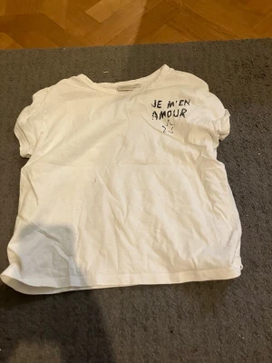 Vit t-shirt med tryck och stjärna - Vit t-shirt med svart texten 'JE M'EN AMOUR' och en stjärna tryckt både fram och bak. T-shirten har korta ärmar och en avslappnad passform. Perfekt för en chill och enkel stil.